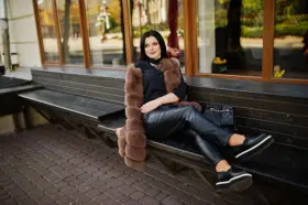 Elegancka kobieta w futrzanej kamizelce i skórzanych spodniach, zastanawia się, jakie buty do skorzanych spodni będą najlepsze. Wybrała czarne, platformowe mokasyny.