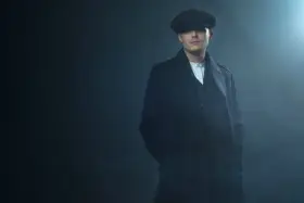 Mężczyzna w kaszkiecie i płaszczu, z charakterystycznym wąsem, emanuje chłodnym spojrzeniem w stylu Peaky Blinders.