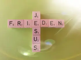 Buchstabensteine bilden ein Kreuz mit den Wörtern "FRIEDEN" und "JESUS". Ein Zitat aus Johannes 14.