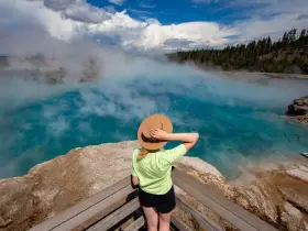 Kobieta w kapeluszu podziwia turkusowe gorące źródło w Yellowstone. Gdzie jest Yellowstone? Tu, wśród parujących wód i lasów.