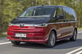 Nowy Volkswagen van w kolorze czerwono-czarnym pędzi po drodze, otoczony zielenią drzew.