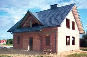 Budowa domu z cegły, widoczny dach betonowy skośny ocieplenie. Widać otwory okienne i drzwiowe, komin.