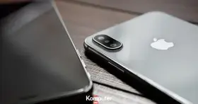 Dwa telefony leżą obok siebie na drewnianym blacie. Jeden z nich, srebrny iPhone, ma widoczny moduł aparatu. Drugi, z ciemnym ekranem, jest ustawiony bokiem. Idealne do rozmowy przez **Facetime**.