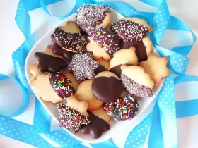 Un plato lleno de pastas de té caseras, algunas cubiertas de chocolate y chispas de colores, otras con coco rallado, rodeadas de cintas azules.