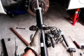 Mano guantata tiene un ammortizzatore nuovo, pronto per sostituire quelli scarichi dell'auto. Attrezzi e molle a vista.