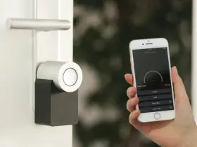 Mano sostiene un iPhone que muestra una app para controlar un cerrojo inteligente. La puerta tiene un diseño moderno.