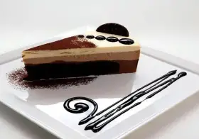 Una rebanada de tarta de tres chocolates, con capas de chocolate blanco, con leche y negro, decorada con cacao en polvo y un disco de chocolate.