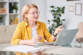 Kobieta w okularach i żółtej koszuli pracuje przy laptopie, trzymając długopis. To jej dodatek funkcyjny do pracy zdalnej.