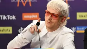 Joost Klein w białej koszuli i różowych okularach przeciwsłonecznych, z charakterystyczną fryzurą, wskazuje palcem w stronę mikrofonu.