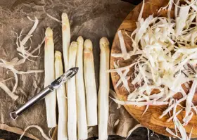 Frischer weißer Spargel wird geschält, die Spargelschalen liegen daneben. Ein Sparschäler liegt auf den Spargelstangen.