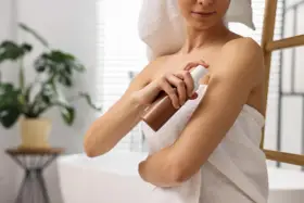 Kobieta aplikuje samoopalacz na ramię, dbając o równomierny koloryt skóry, nawet wokół tatuażu.