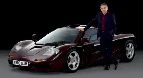 Rowan Atkinson w garniturze opiera się o swój bordowy McLaren F1. To jeden z jego ulubionych samochodów.