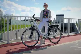 Kobieta w białych spodniach i kolorowej koszuli stoi obok białego roweru elektrycznego. Czy na rower elektryczny trzeba mieć prawo jazdy?