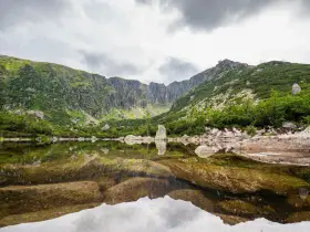 Spokojne, górskie jezioro odbija skaliste zbocza i chmury. W oddali widać skalne formacje, a brzegi porasta bujna zieleń. To idealne miejsce na podziwianie piękna przyrody, jakbyśmy patrzyli na śnieżne stawki.