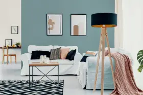 Nowoczesny salon z białymi sofami, stolikiem kawowym i lampą na drewnianym statywie. Ściana w odcieniu **kolor lazurowy** tworzy spokojną atmosferę.