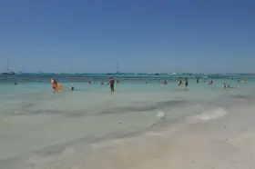 Słoneczny dzień na plaży Es Trenc. Ludzie kąpią się w turkusowej wodzie, a na horyzoncie widać żaglówki.