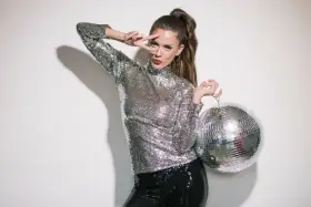 Kobieta w cekinowym topie i spodniach, z kulą dyskotekową w ręku, pozuje w stylu disco.