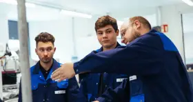 Drei junge Männer in blauen Arbeitskitteln lernen gemeinsam. Ein Ausbilder erklärt etwas, während die genz-Auszubildenden aufmerksam zuhören.