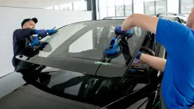 Dwóch mechaników wymienia przednią **szybę** w czarnym samochodzie, używając przyssawek.