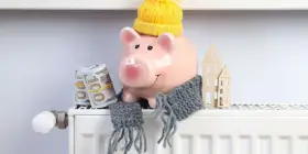 Świnka-skarbonka w czapce i szaliku, z banknotami i domkami na grzejniku. Oszczędzanie na rachunki za ogrzewanie, czyli bon ciepłowniczy w praktyce.