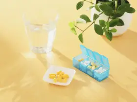 Un bicchiere d'acqua, una ciotolina con capsule gialle e un portapillole blu. Forse integratori per capire cosa sono gli elettroliti.