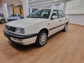Biały Volkswagen Vento z lat 90. stoi na drewnianej podłodze w salonie samochodowym.