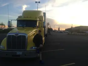 Młody mężczyzna opiera się o żółtą ciężarówkę. W tle zachód słońca i parking pełen aut. To obrazek z życia polskie truckerki.