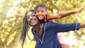 Une femme porte une enfant sur son dos, toutes deux souriantes, avec des arbres en arrière-plan.