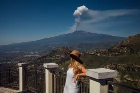 Kobieta w kapeluszu patrzy na dymiący wulkan Etna z tarasu widokowego.