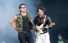 Albert Hammond Jr. w kamizelce moro i okularach stoi obok gitarzysty w skórzanej kurtce, grającego na białej gitarze.