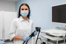 Lekarka w masce i rękawiczkach trzyma endoskop, przygotowując się do gastroskopii. Obok monitor i sprzęt medyczny.