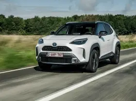 Biały Yaris Cross Hybrid jedzie po drodze, otoczony zielenią.
