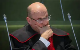 Sędzia w todze z czerwoną lamówką, w okularach, z ręką na brodzie, zamyślony.