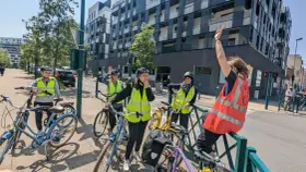 Des adultes souriants, casqués et en gilets fluo, prêts à apprendre à faire du vélo adulte. Ils sont avec leurs vélos, devant un immeuble moderne.