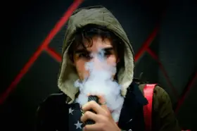 Joven con capucha exhala vapor de un vapeador, con un diseño de estrellas en su camiseta. Los nicokits son su secreto.