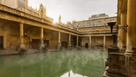 Steaming green water fills the Great Bath, surrounded by ancient Roman ruins with columns and torches.