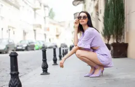 Kobieta w liliowej sukience i pasujących do niej butach na obcasie, siedzi na chodniku. Idealne buty do liliowej sukienki.