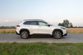 Biała Toyota Corolla Cross Hybrid stoi na poboczu drogi, otoczona polami i zieloną trawą.