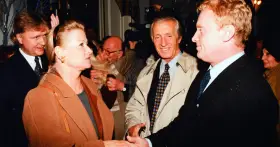 Monika Dzienisiewicz-Olbrychska w brązowym płaszczu wita się z mężczyznami. W tle widać kamery i innych ludzi.
