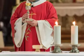 Kapłan w czerwonej szacie z krzyżem trzyma kielich podczas konsekracji. Płoną dwie świece.