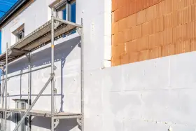 Budynek w trakcie ocieplania. Widoczna fasada z białym styropianem i fragmentem cegły klinkierowej. Rusztowanie ułatwia pracę.