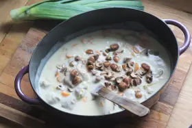 Un plat de blanquette de veau facile mijote dans une cocotte, garni de champignons et de carottes. Un poireau est posé à côté.