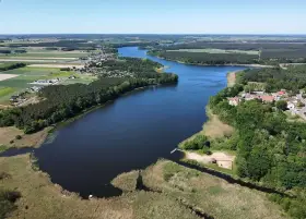 Malownicze jezioro Wojnowskie otoczone lasami i polami. W oddali widać zabudowania wsi, a na brzegu plażę z pomostem.