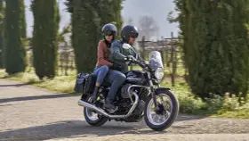 Coppia su una Moto Guzzi V7, con passeggero, percorre una strada alberata in una giornata di sole.