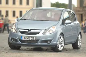 Jasnoniebieski Opel Corsa D z włączonymi żarówkami z przodu, zaparkowany na bruku.