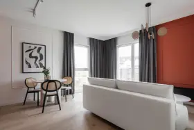 Jasny salon z białym narożnikiem, okrągłym stolikiem i krzesłami z rattanu. Ściana w kolorze ceglastym. Standardowa wysokość mieszkania.