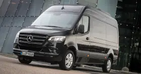 Czarny furgon Mercedes-Benz Sprinter stoi przed nowoczesnym budynkiem z przeszkloną fasadą.
