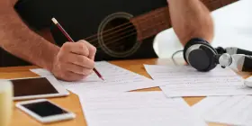En person skriver noter med en penna, med en gitarr och hörlurar i bakgrunden. Detta är en bra inledning till en ny låt.
