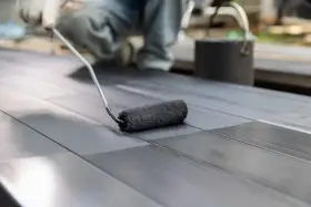 Osoba w rękawiczkach maluje wałkiem metalowe profile. Powierzchnia jest ciemna, prawdopodobnie zabezpieczana przed korozją, jak elastometal.