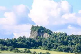 Skalisty szczyt porośnięty drzewami, przypominający wilczą górę w Złotoryi, góruje nad zielonymi polami i lasami.